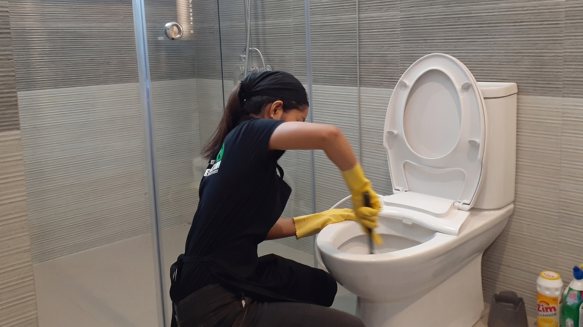 A CleanX employee wearing yellow rubber gloves scrubbing the inside of a bathroom toilet bowl during a deep cleaning service.
