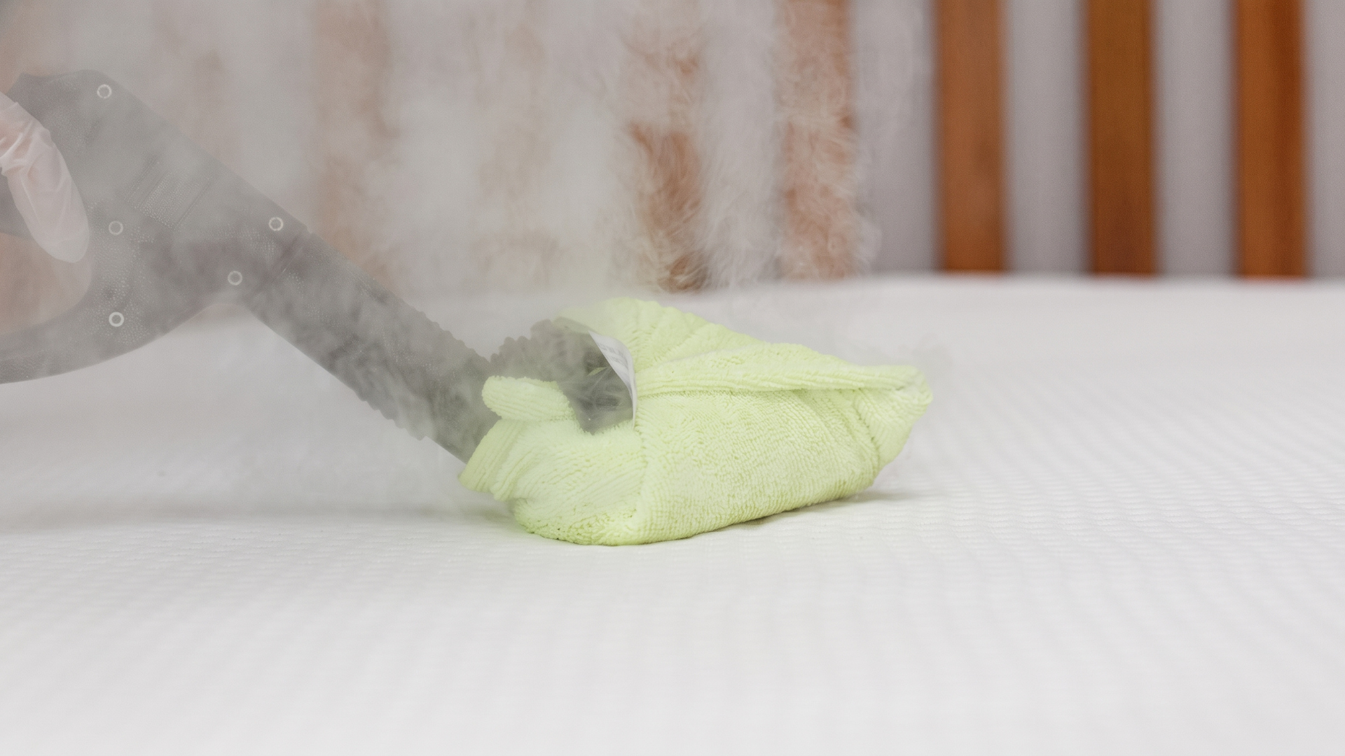 Close-up of a CleanX steam cleaning tool wrapped in a green microfiber cloth emitting hot steam to sanitize a white mattress.