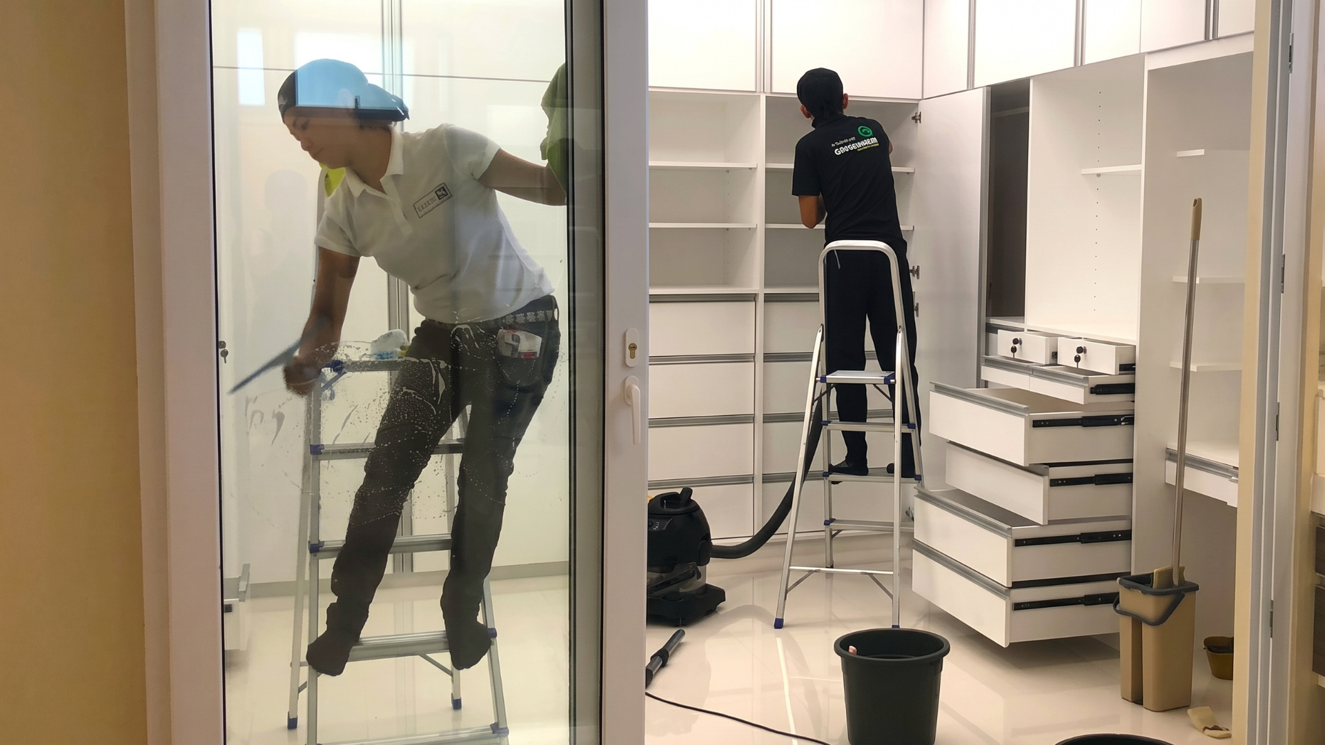 Two CleanX staff members working in a room; one is squeegeeing a glass door while the other stands on a ladder vacuuming inside a large white custom closet.
